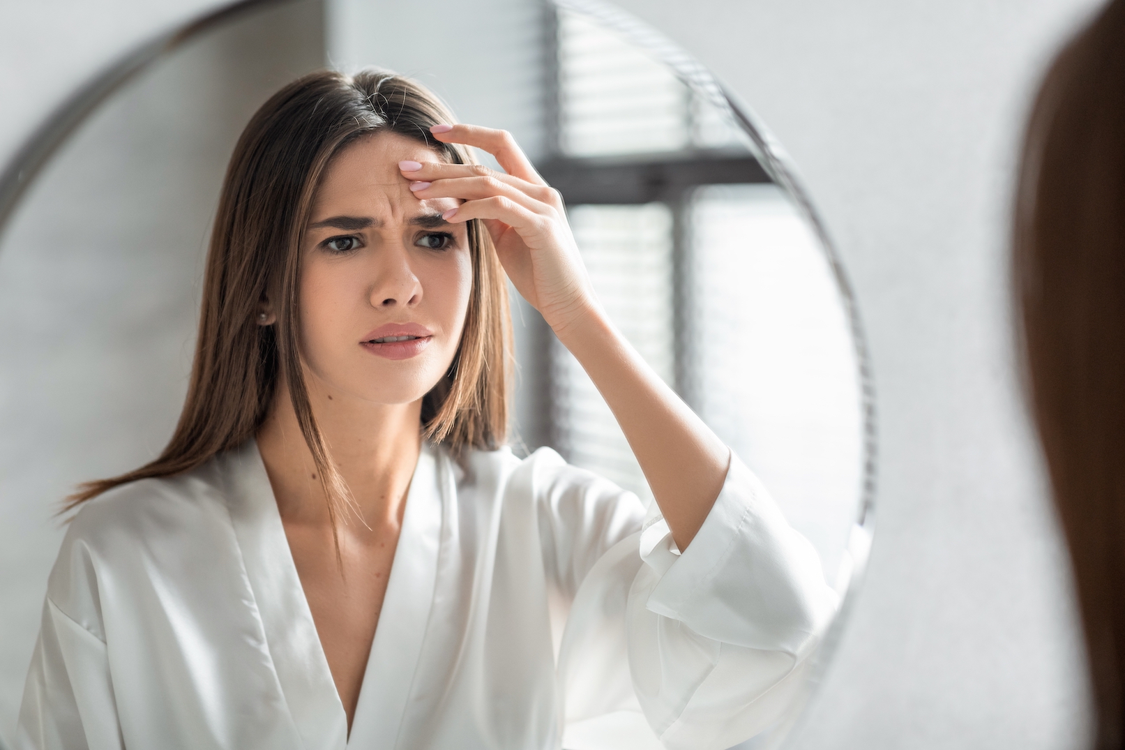 Unhappy Young Woman Looking In Mirror And Touching Wrinkles On Her Face, Attractive Millennial Female Standing In Bathroom And Examining Skin Fine Lines, Selective Focus On Reflection, Closeup